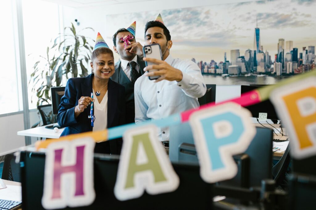 Three colleagues celebrating a birthday in the office with party hats and decorations, capturing a fun selfie.